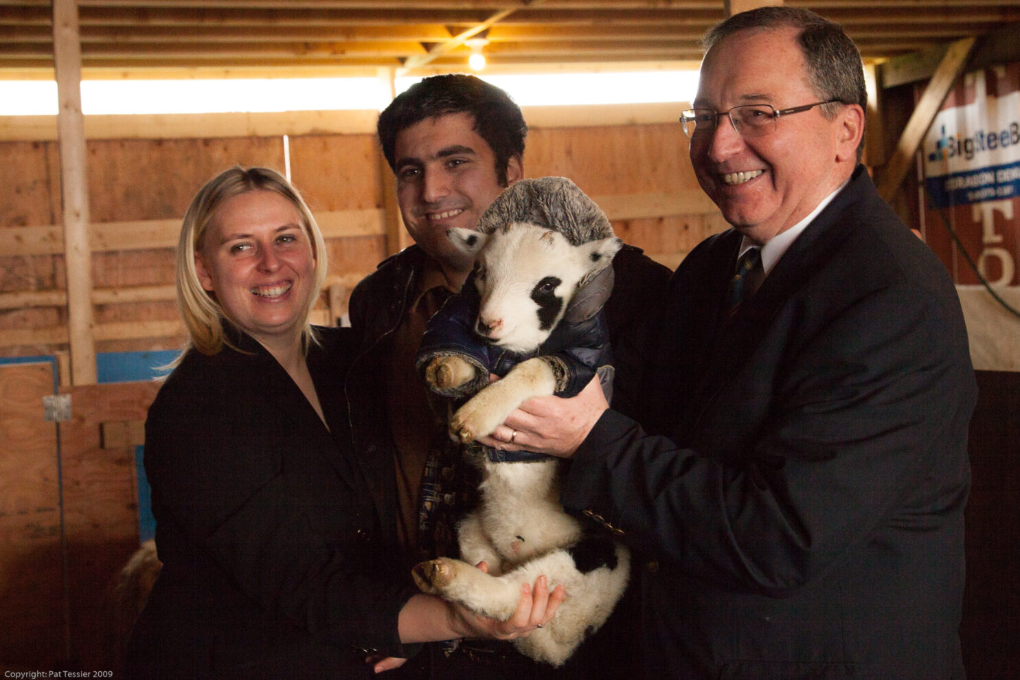 Three adults—a woman with blonde hair on the left, a man with dark hair in the middle, and a man wearing glasses and a dark suit on the right—are smiling as they jointly hold a small Jacob Sheep lamb in an indoor barn setting with wooden walls. The lamb is white with black patches on its face and body, and it is wearing a small blue coat.