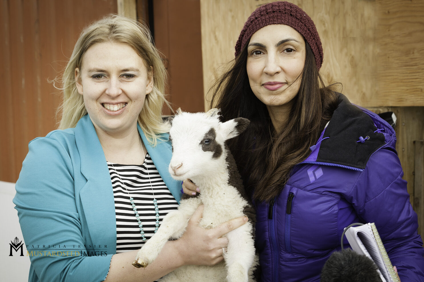 Two women are posing for a photo outdoors, standing in front of a blurred reddish-brown wooden wall. The woman on the left, wearing a light blue blazer over a striped shirt, is holding a small, white lamb with black patches around its eyes and ears. The woman on the right is wearing a dark beanie and a blue puffer jacket, looking towards the camera.