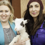 Two women are posing for a photo outdoors, standing in front of a blurred reddish-brown wooden wall. The woman on the left, wearing a light blue blazer over a striped shirt, is holding a small, white lamb with black patches around its eyes and ears. The woman on the right is wearing a dark beanie and a blue puffer jacket, looking towards the camera.