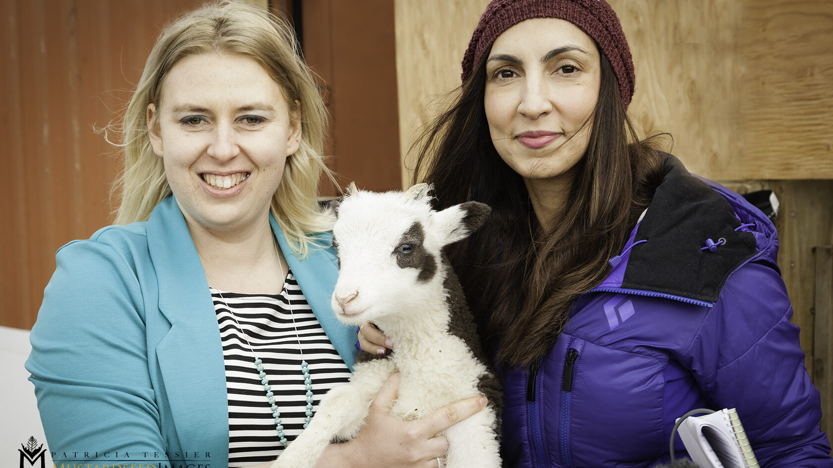 Two women are posing for a photo outdoors, standing in front of a blurred reddish-brown wooden wall. The woman on the left, wearing a light blue blazer over a striped shirt, is holding a small, white lamb with black patches around its eyes and ears. The woman on the right is wearing a dark beanie and a blue puffer jacket, looking towards the camera.