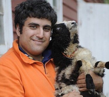 A man with dark, curly hair and a beard, wearing a bright orange long-sleeved shirt, is smiling while holding a small Jacob Sheep lamb. The lamb is white with distinct black markings on its face and body, and wears a small light-colored beaded collar. It is looking upwards. They are outdoors in front of a blurred rustic building with red and white painted walls.