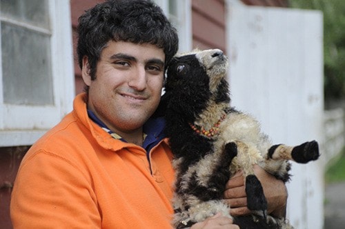 A man with dark, curly hair and a beard, wearing a bright orange long-sleeved shirt, is smiling while holding a small Jacob Sheep lamb. The lamb is white with distinct black markings on its face and body, and wears a small light-colored beaded collar. It is looking upwards. They are outdoors in front of a blurred rustic building with red and white painted walls.