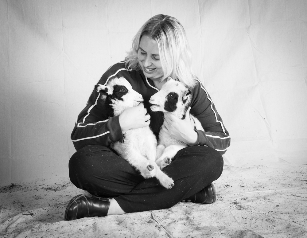 In a black and white photograph, a woman with blonde hair sits cross-legged on a light-colored surface. She is wearing dark, long-sleeved clothing with light stripes on the sleeves and is gently holding two small Jacob Sheep lambs in her arms. Both lambs are white with distinct black patches around their eyes and ears, facing each other. The background is a plain, light-toned backdrop.