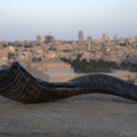 A dark, curved shofar (ram's horn) rests horizontally on a rough, light-colored stone ledge in the foreground. In the blurred background, the city of Jerusalem stretches out under a twilight sky, with the distinctive golden dome of the Dome of the Rock visible in the mid-ground among other buildings and distant structures.