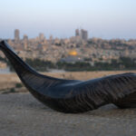 A dark, curved shofar with visible texture rests horizontally on a weathered stone surface in the foreground, filling the lower portion of the frame. In the blurred background, the iconic cityscape of Jerusalem is visible under a pale sky, prominently featuring the golden dome of the Dome of the Rock among other ancient and modern buildings.