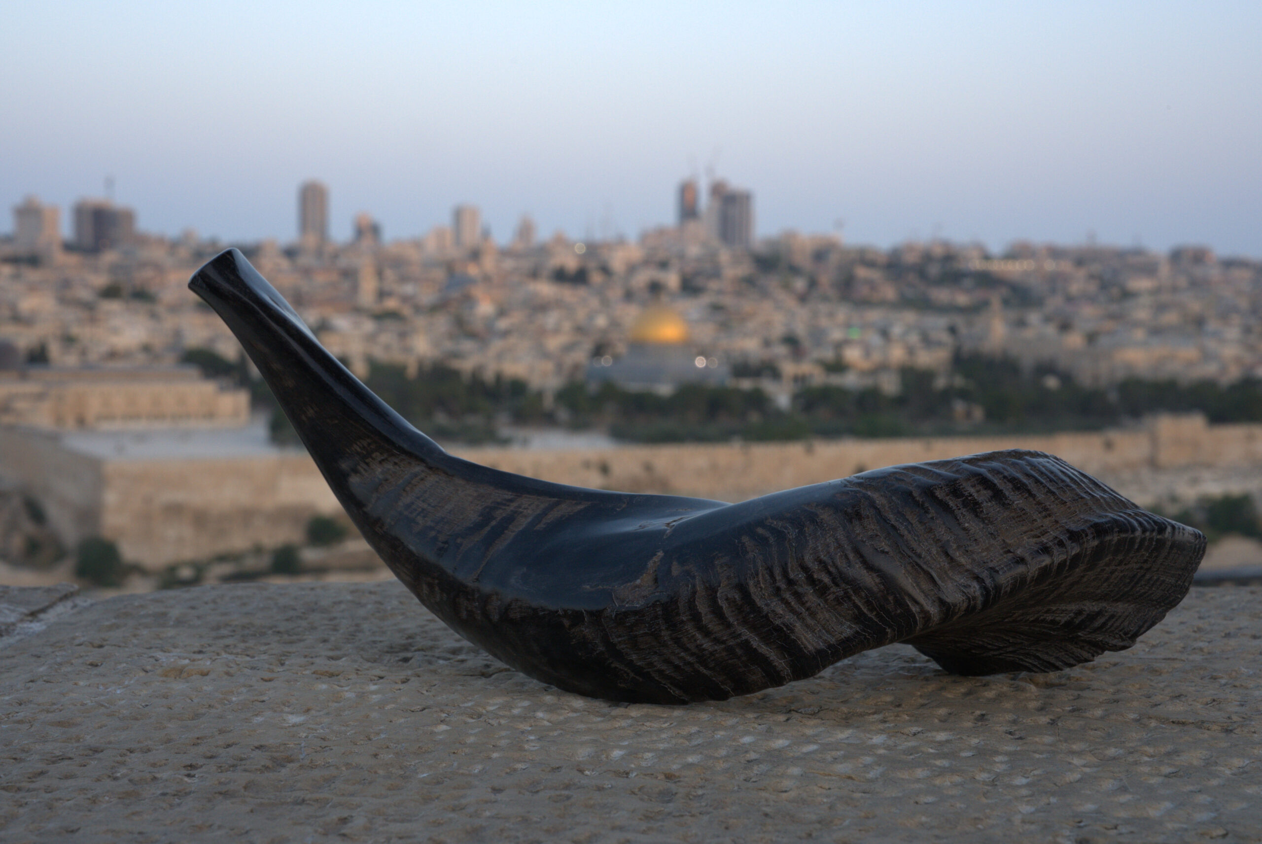 A dark, curved shofar with visible texture rests horizontally on a weathered stone surface in the foreground, filling the lower portion of the frame. In the blurred background, the iconic cityscape of Jerusalem is visible under a pale sky, prominently featuring the golden dome of the Dome of the Rock among other ancient and modern buildings.
