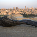 A dark, highly curved shofar with prominent horizontal ridges and a slight sheen rests on a light-colored, textured stone surface. The shofar's wide, open end on the left shows a deep internal curve. In the blurred background, the cityscape of Jerusalem, including its golden-domed landmark, is bathed in the soft glow of dusk or dawn beneath a pale sky.