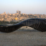 A dark, highly curved shofar with fine, horizontal striations along its body rests on a light-colored, granular stone surface. The shofar's wide opening, appearing round and dark, is visible on the left. In the blurred background, the iconic Jerusalem skyline, including the golden Dome of the Rock, is silhouetted against a pale, evening sky.