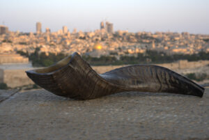 A dark, polished shofar with textured ridges is positioned horizontally on a light-colored, granular stone surface in the foreground. Its wider, opening end is to the left. In the softly blurred background, the cityscape of Jerusalem unfolds under a pale, twilight sky, with the golden dome of the Dome of the Rock distinct in the mid-ground among clusters of buildings.
