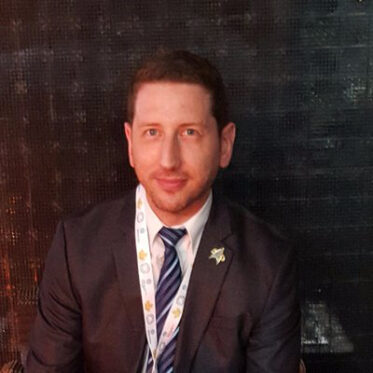 A headshot portrait of an adult man with short, light brown hair and a beard, looking directly at the camera with a slight smile. He is wearing a dark suit jacket, a white collared shirt, and a blue and white striped tie. A lanyard with a patterned ribbon is around his neck, and a small pin is on his lapel. The background is dark and blurred.