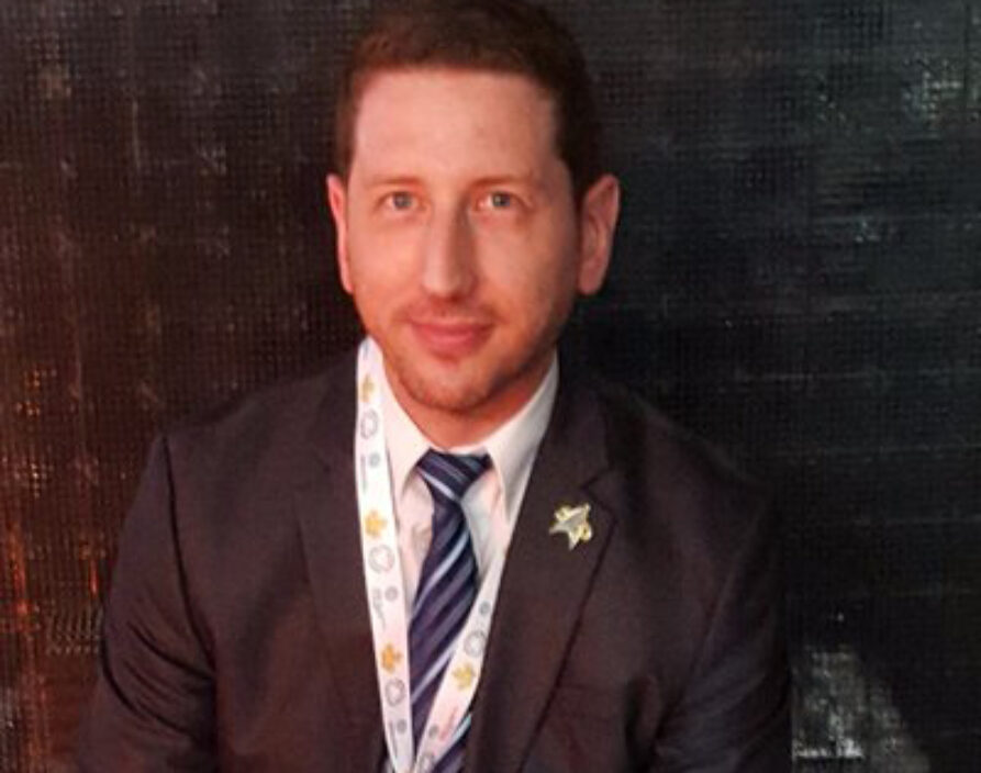 A headshot portrait of an adult man with short, light brown hair and a beard, looking directly at the camera with a slight smile. He is wearing a dark suit jacket, a white collared shirt, and a blue and white striped tie. A lanyard with a patterned ribbon is around his neck, and a small pin is on his lapel. The background is dark and blurred.
