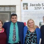 Four adults, two men and two women, stand smiling directly at the camera. Behind them is a building and a large white informational sign with the words "Jacob Sheep" in English and Hebrew. The man second from the left is wearing a kippah. All individuals are dressed in business or formal attire.