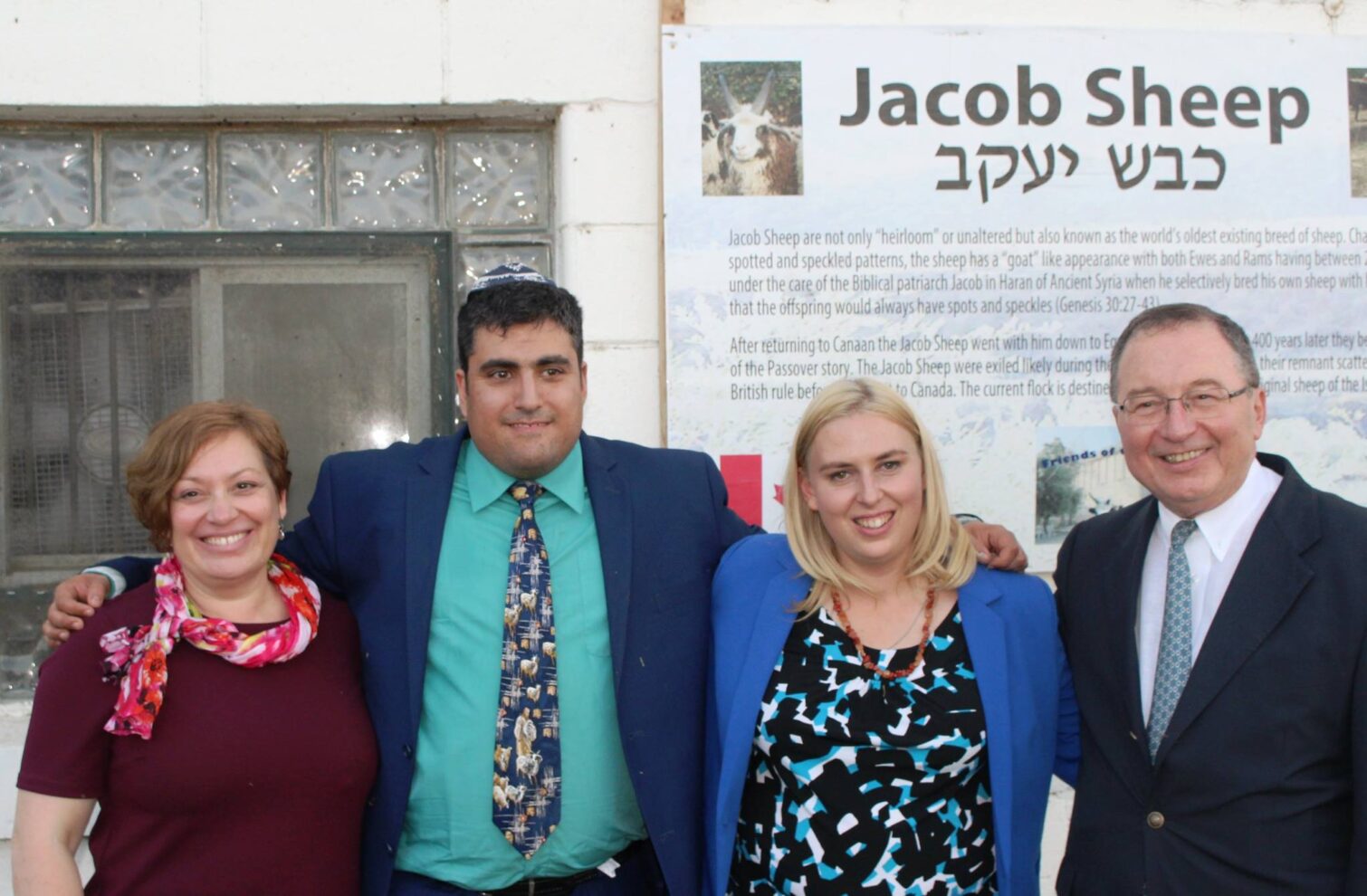 Four adults, two men and two women, stand smiling directly at the camera. Behind them is a building and a large white informational sign with the words "Jacob Sheep" in English and Hebrew. The man second from the left is wearing a kippah. All individuals are dressed in business or formal attire.