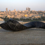 A dark, highly curved shofar with a textured, polished surface rests horizontally on a light-colored, mottled stone ledge in the foreground. The shofar's wide end is subtly open to the left. Behind it, the blurred cityscape of Jerusalem unfolds under a pale sky, prominently featuring the golden dome of the Dome of the Rock and other buildings, conveying depth of field.