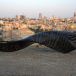 A dark-toned shofar, with a smooth, polished finish contrasting with subtle horizontal striations, rests horizontally on a textured, light grey stone surface in the foreground. Its wider, open end points to the left. In the blurred background, the iconic city of Jerusalem is illuminated by the soft light of dusk or dawn, with its golden-domed landmark distinct amidst countless buildings.