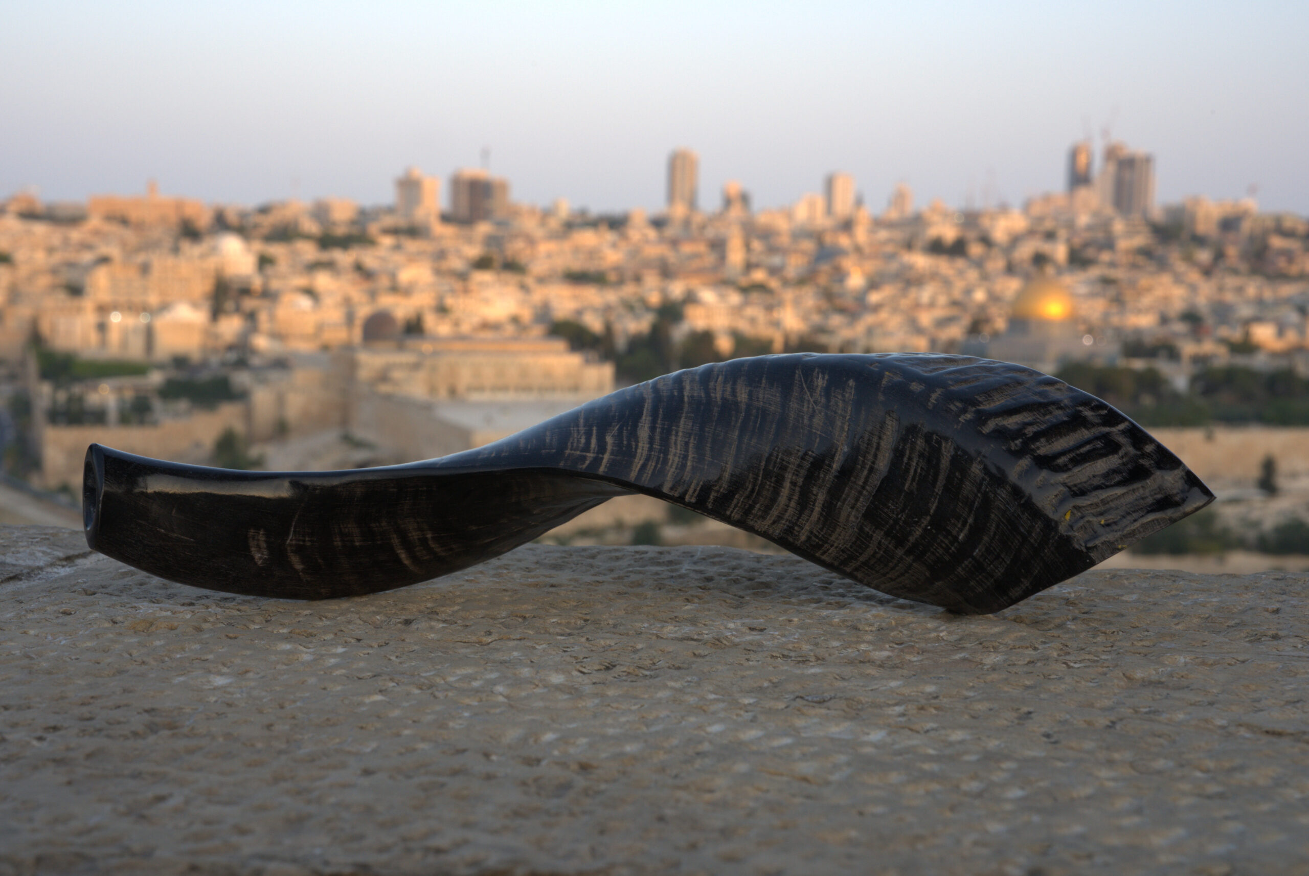 A dark-toned shofar, with a smooth, polished finish contrasting with subtle horizontal striations, rests horizontally on a textured, light grey stone surface in the foreground. Its wider, open end points to the left. In the blurred background, the iconic city of Jerusalem is illuminated by the soft light of dusk or dawn, with its golden-domed landmark distinct amidst countless buildings.