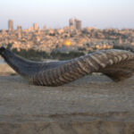 A long, curved shofar with prominent, wide horizontal bands of light and dark grey-brown colors rests on a rough, light-toned stone surface in the foreground. Its wide, open end is on the left. In the blurred background, the panoramic cityscape of Jerusalem is visible under a pale, clear sky, featuring the golden dome of the Dome of the Rock among various buildings.