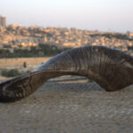 A dark, textured shofar with prominent horizontal ridges rests on a light-colored, pebbled stone surface in the foreground. The shofar's narrower, open end is on the left, curving upwards. In the blurred background, the panoramic cityscape of Jerusalem is visible, featuring the golden Dome of the Rock amidst countless buildings under a pale, diffused sky.