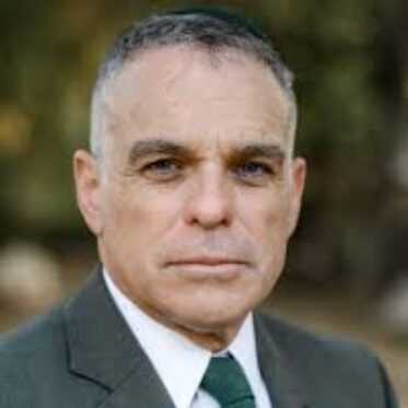 A headshot portrait of an adult man with short, graying hair, looking directly at the camera with a neutral expression. He is wearing a dark suit jacket, a white collared shirt, a dark green tie, and a small, dark circular head covering. The background is a blurred outdoor scene of green and brown foliage.