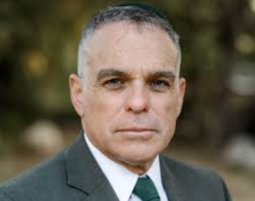 A headshot portrait of an adult man with short, graying hair, looking directly at the camera with a neutral expression. He is wearing a dark suit jacket, a white collared shirt, a dark green tie, and a small, dark circular head covering. The background is a blurred outdoor scene of green and brown foliage.