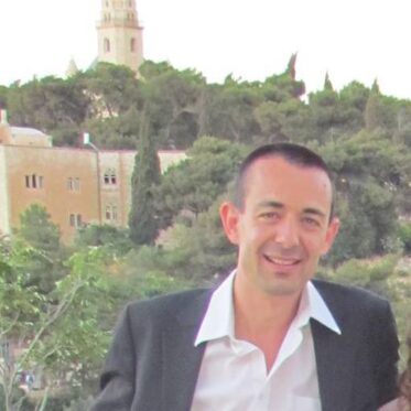 A man with short, dark hair and a broad smile is seen from the chest up, wearing a dark blazer over a white collared shirt with an open collar. The background is a blurred outdoor landscape featuring dense green trees and a large, light-colored building with a prominent tower.