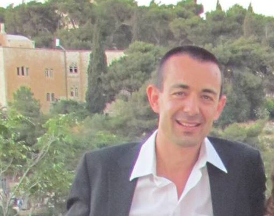 A man with short, dark hair and a broad smile is seen from the chest up, wearing a dark blazer over a white collared shirt with an open collar. The background is a blurred outdoor landscape featuring dense green trees and a large, light-colored building with a prominent tower.