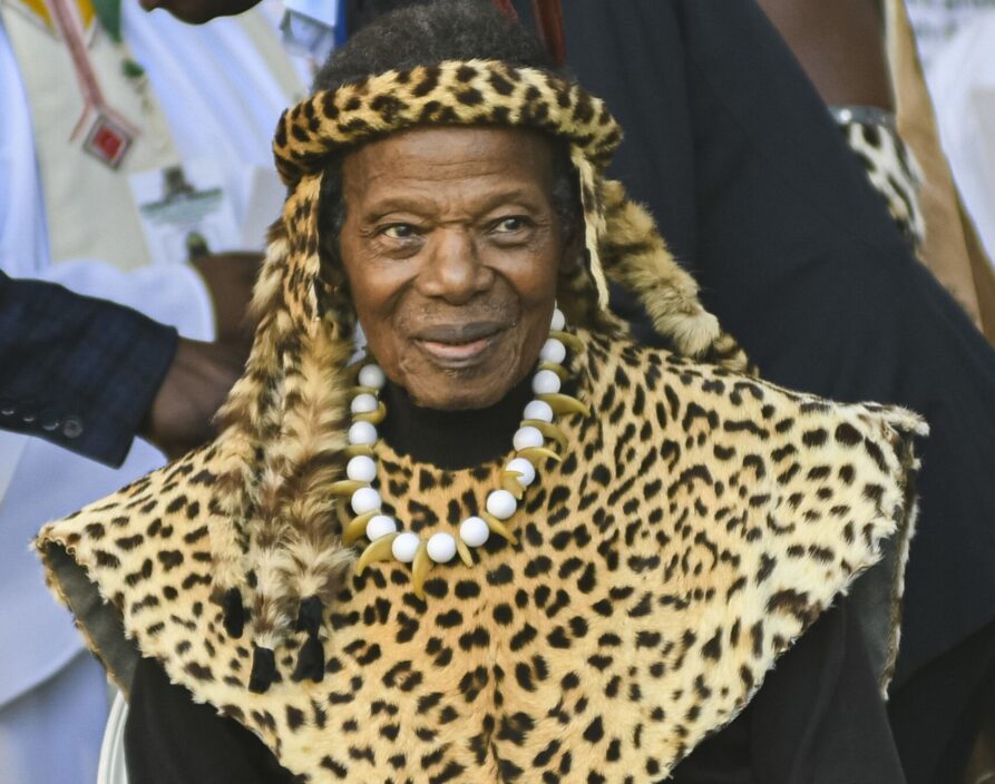An elderly person is seated, wearing an elaborate outfit featuring a leopard-print mantle with a matching headband. They are adorned with a multi-strand white beaded necklace and large light-colored wrist cuffs. Their lower legs have thick, white, furry wraps around the ankles, and they hold a thin staff. The background is blurred, with other people partially visible.