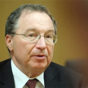 A headshot portrait of an adult man with short, graying hair and glasses, wearing a dark suit jacket, white collared shirt, and a reddish patterned tie. His mouth is slightly open, and he is looking slightly off to the right. The background is a blurred, solid brown.