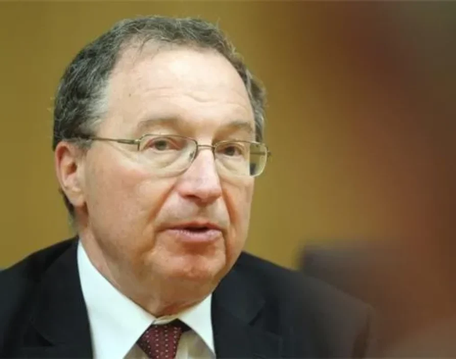 A headshot portrait of an adult man with short, graying hair and glasses, wearing a dark suit jacket, white collared shirt, and a reddish patterned tie. His mouth is slightly open, and he is looking slightly off to the right. The background is a blurred, solid brown.