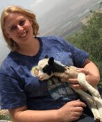 A blonde woman in a blue patterned t-shirt is smiling directly at the camera while gently holding a small Jacob Sheep lamb. The lamb is white with black markings around its eyes and ears, and is resting comfortably in her arms. Behind them is an expansive, blurred outdoor landscape of rolling hills and a valley under a bright, hazy sky, with some green foliage in the immediate foreground.