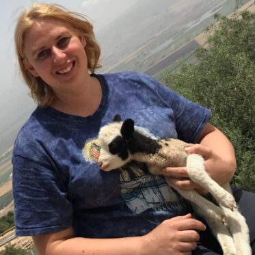 A blonde woman in a blue patterned t-shirt is smiling directly at the camera while gently holding a small Jacob Sheep lamb. The lamb is white with black markings around its eyes and ears, and is resting comfortably in her arms. Behind them is an expansive, blurred outdoor landscape of rolling hills and a valley under a bright, hazy sky, with some green foliage in the immediate foreground.