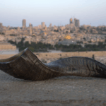 A dark, curved shofar (ram's horn) rests horizontally on a rough, light-colored stone ledge in the foreground. In the blurred background, the city of Jerusalem stretches out under a twilight sky, with the distinctive golden dome of the Dome of the Rock visible in the mid-ground among other buildings and distant structures.