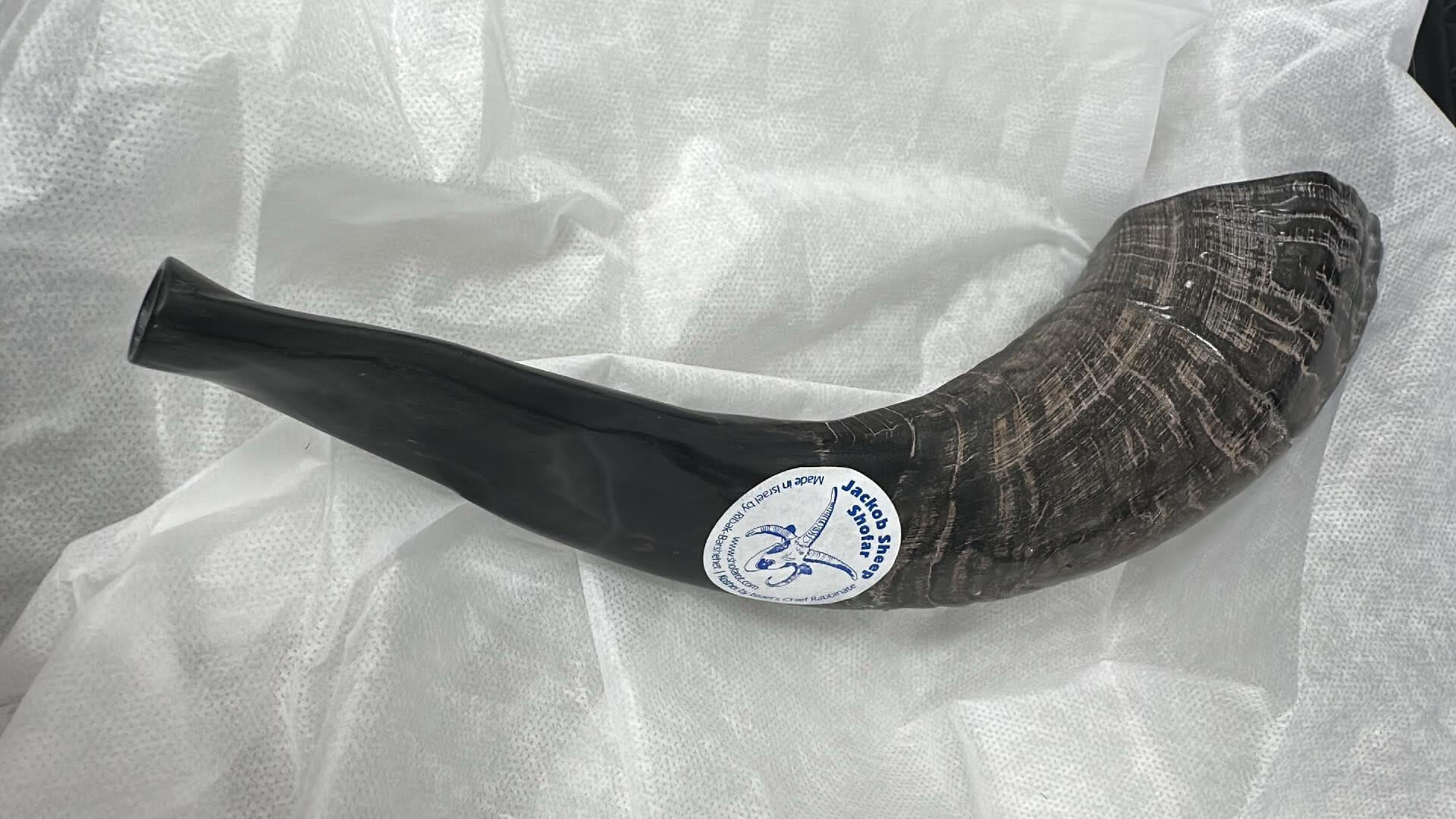 A dark, curved shofar (ram's horn) with visible ridges lies on a crumpled white fabric surface. A round white sticker with a blue border is affixed to its side, clearly displaying an illustration of a sheep's head and the text "Jacob Sheep Shofar".