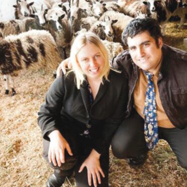 A blonde woman in a dark blazer and a man in a dark jacket with a blue patterned tie are kneeling on hay-covered ground, smiling towards the camera. Behind them, a large flock of Jacob Sheep, displaying various black, white, and brown wool patterns and horns, fills the background of a barn. A sheep with distinct vertical stripes is visible on the far left.